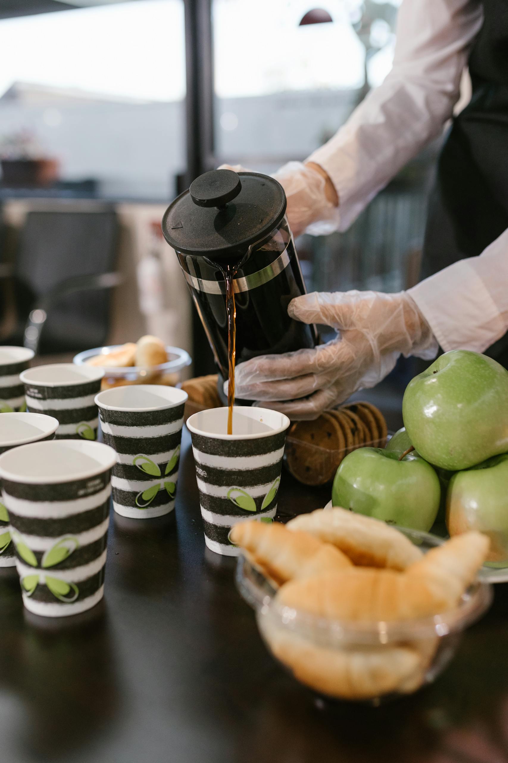 Barista pours coffee from French press into cups at a breakfast event with croissants and apples.